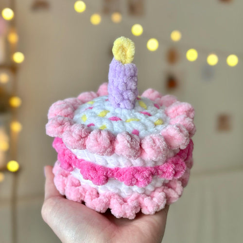 Pink pom-pom birthday cake with a candle held in front of a softly blurred background with lights.
